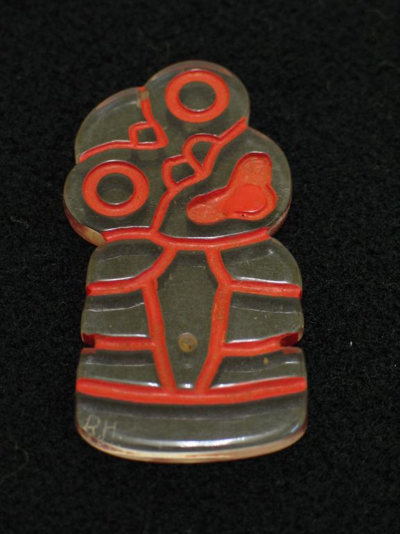 2010-067.14 Tiki pendant made from clear plexiglass (perspex). Carved details have been highlighted with red paint. Made while with Royal New Zealand Air Force No. 12 Servicing Unit at Henderson Field, Guadalcanal.