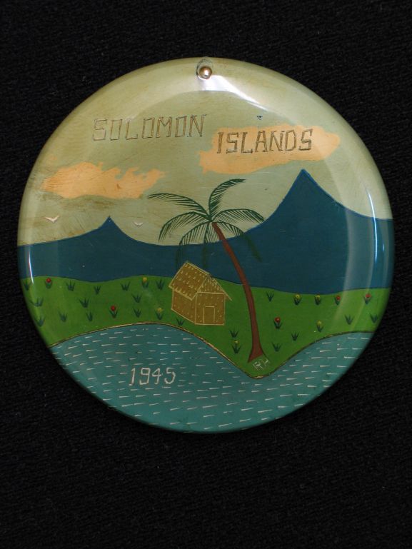 2010-067.15 Round plexiglass pendant engraved with island beach scene - water, beach, hut, palm tree and mountains. "SOLOMON ISLANDS " engraved above mountains and "1945" in middle of water. Engraving has been highlighted by the reverse of pendant being painted. Made while with Royal New Zealand Air Force No. 12 Servicing Unit at Henderson Field, Guadalcanal.